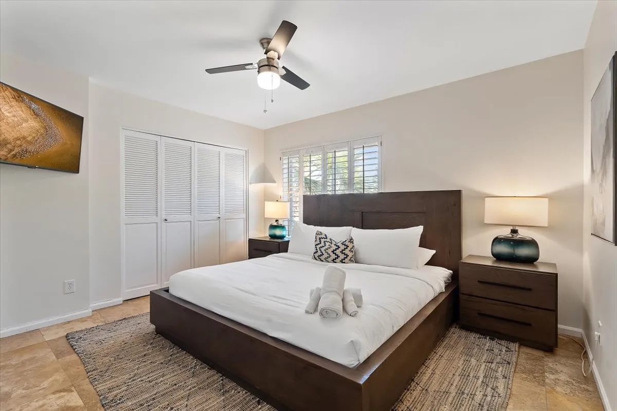 A spacious king bedroom with a large bed, fluffy bedding, matching nightstands with lamps, and a television mounted on the wall. Natural light streams through a window, creating an airy and inviting atmosphere ideal for unwinding after a busy day.