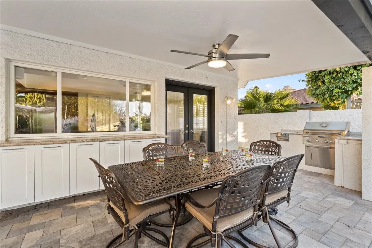 This exterior scene captures a stylish outdoor dining area with a large table, grill, palm trees, and expansive windows connecting indoors and outdoors. The setup suggests a space perfect for al fresco meals, barbecues, and relaxed social gatherings after a productive workday.