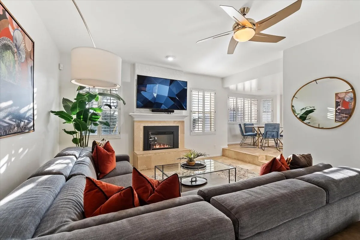 A cozy, modern living room with a sofa adorned with red pillows, a stylish fireplace, a large television, and a dining area visible in the background. The ambiance is warm and inviting, perfect for socializing or relaxing.