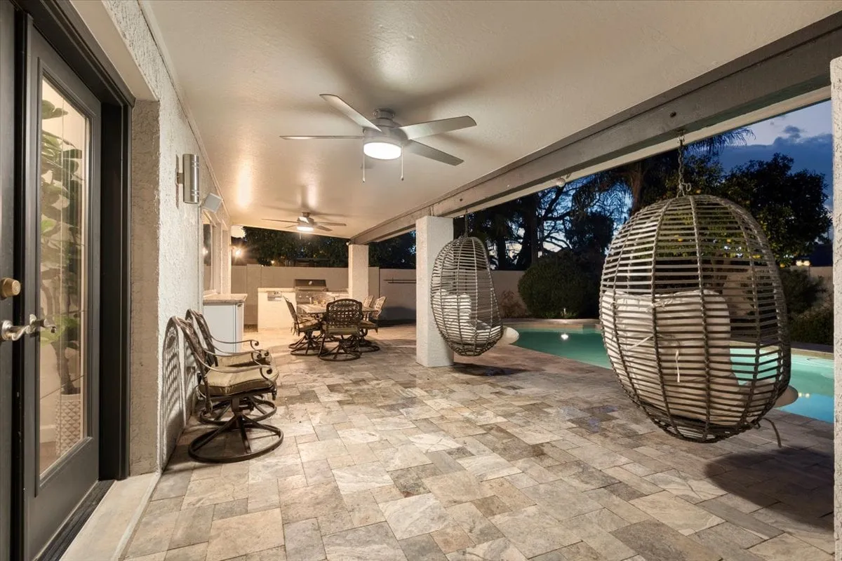An exterior view focusing on hanging chairs, a comfortable outdoor seating area, a sparkling swimming pool, and an outdoor kitchen/BBQ setup. The mood is casual and welcoming, perfect for gatherings or relaxing by the water.