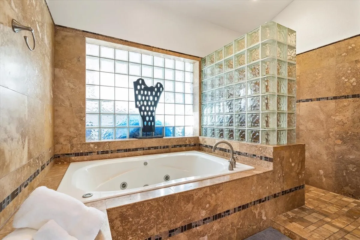 A spa-like bathroom featuring a large jacuzzi tub, glass block wall, and a decorative sculpture. The tranquil ambiance is enhanced by soft lighting and elegant fixtures, evoking the feeling of a personal spa within the home.