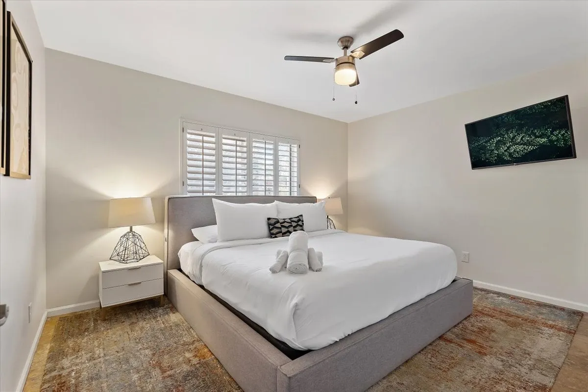 A serene bedroom with plush bedding, decorative pillows, a nightstand with a lamp, and a wall-mounted TV. The room is softly lit, exuding comfort and calmness, perfect for relaxing or reading. The bed is inviting, and the overall decor is modern yet cozy, emphasizing a restful retreat.